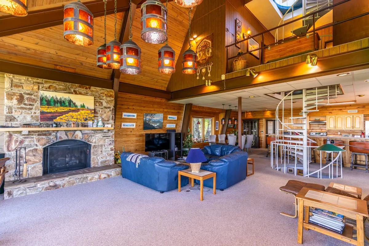 A spacious open-concept area that combines a living room with blue leather sofas, a striking stone fireplace, hanging lanterns, a spiral staircase, and a wooden dining area. The space radiates vintage charm and encourages family gatherings, relaxation, and shared meals, all under abundant natural light.