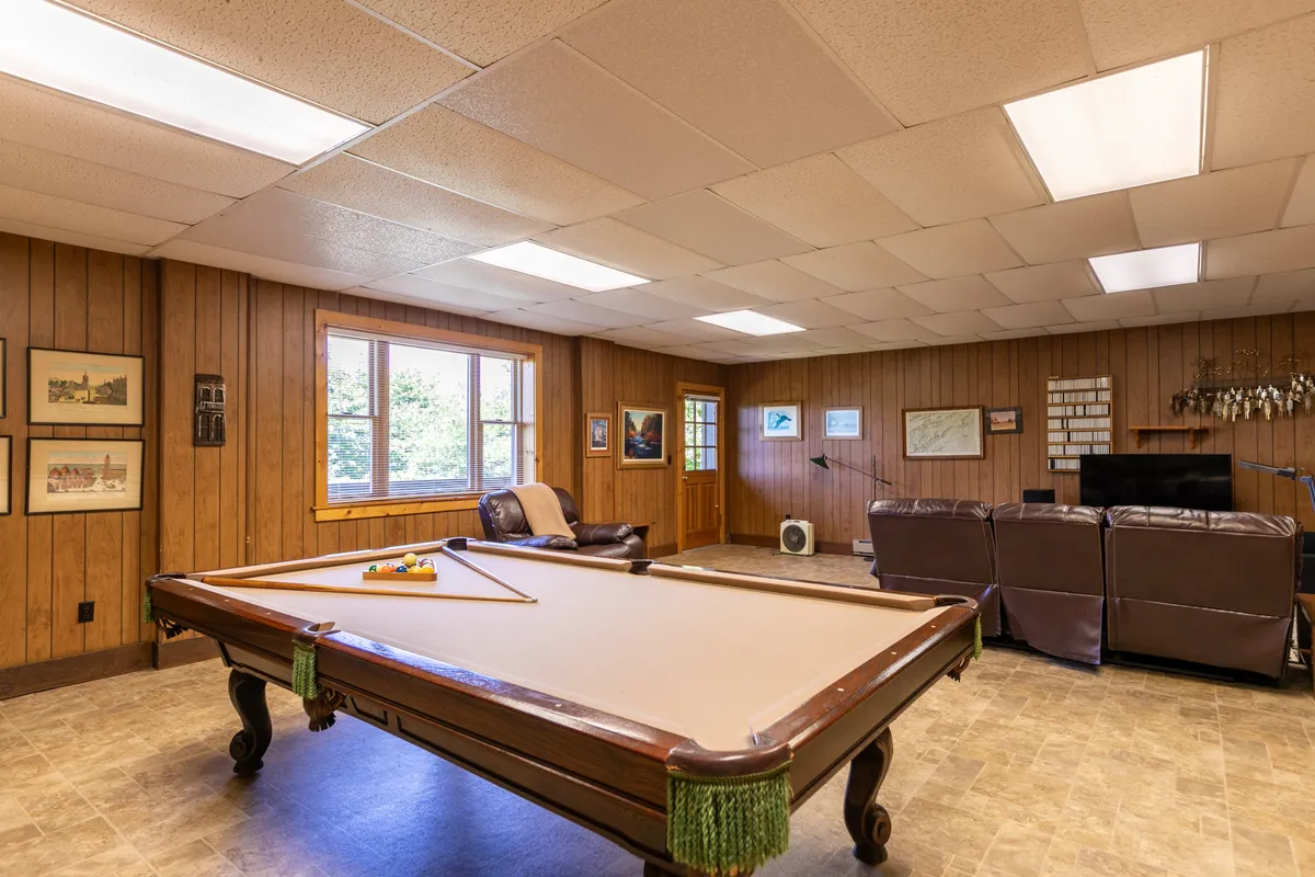 A lively family room featuring a pool table at the center, a plush leather couch facing a large window, and ample natural light filling the space. The room exudes a casual and inviting atmosphere, perfect for fun activities or relaxation.