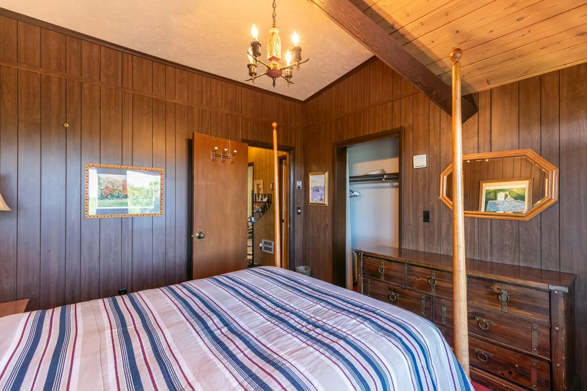 A bedroom with striped bedding, rich wooden paneling, a chandelier above, an antique dresser, and framed artwork. The sunlight filters in, illuminating the cozy and thoughtfully decorated space.