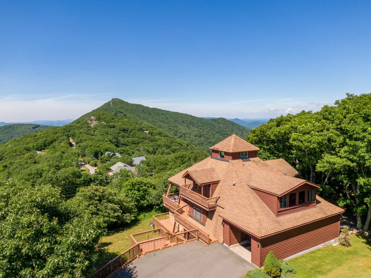 The image showcases a charming mountaintop home with a spacious deck overlooking sweeping mountain vistas. The house sits perched above the surrounding landscape, with the rolling Blue Ridge Mountains stretching out beneath a pastel sky. Comfortable outdoor seating invites you to relax and take in both the sunrise and the sunset, while the yard and natural surroundings hint at peaceful evenings spent stargazing.