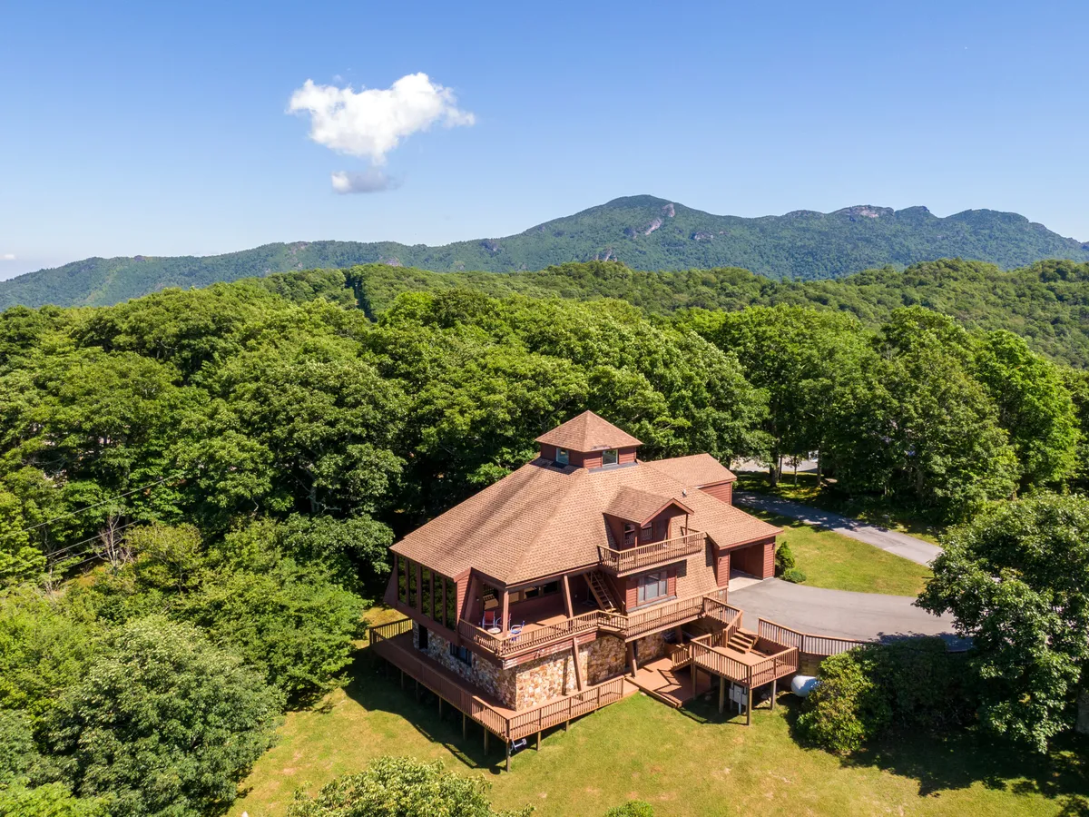 This image features the large wooden house set against a backdrop of lush greenery and mountains. The expansive deck and surrounding lawn area are clearly visible, showcasing ample outdoor space for gatherings, relaxation, and play. The natural setting emphasizes the property's connection to the outdoors and the scenic beauty that guests can enjoy.