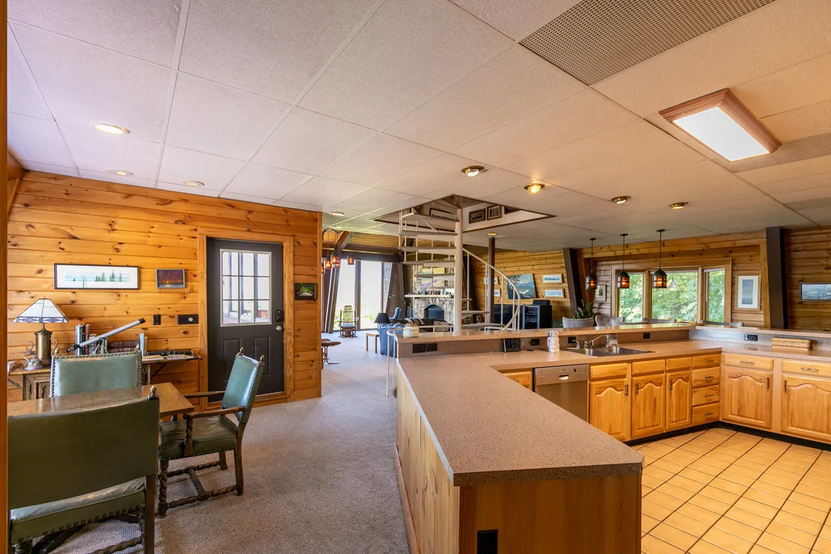 A warm, wood-paneled kitchen and dining area features a central island, spiral staircase, and inviting atmosphere. The space is ideal for hosting family gatherings or holiday dinners, with large windows that likely frame the surrounding natural beauty.