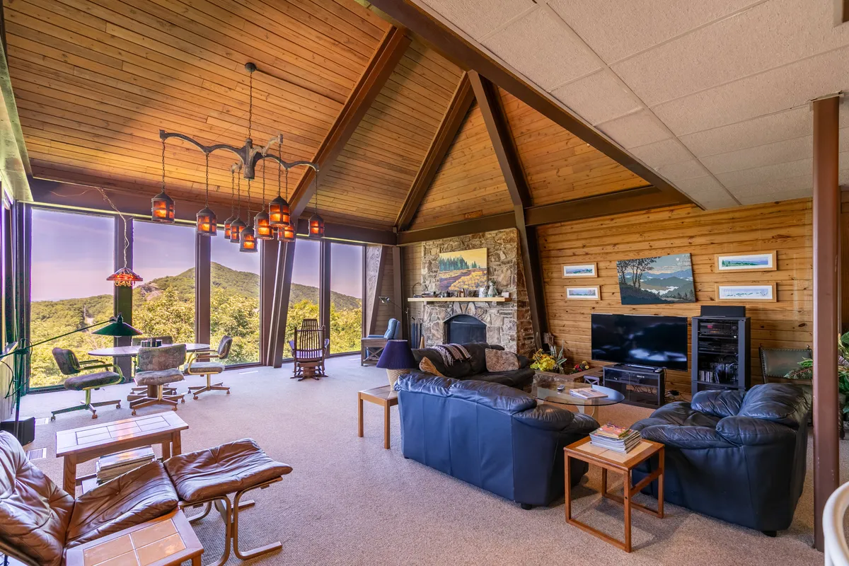 A welcoming living area featuring large, floor-to-ceiling windows that open up to panoramic mountain views. The room is anchored by a dramatic stone fireplace and adorned with exposed wooden beams and comfortable, retro-inspired seating, creating a cozy yet playful atmosphere perfectly in tune with the 70s nostalgia mentioned in the section.
