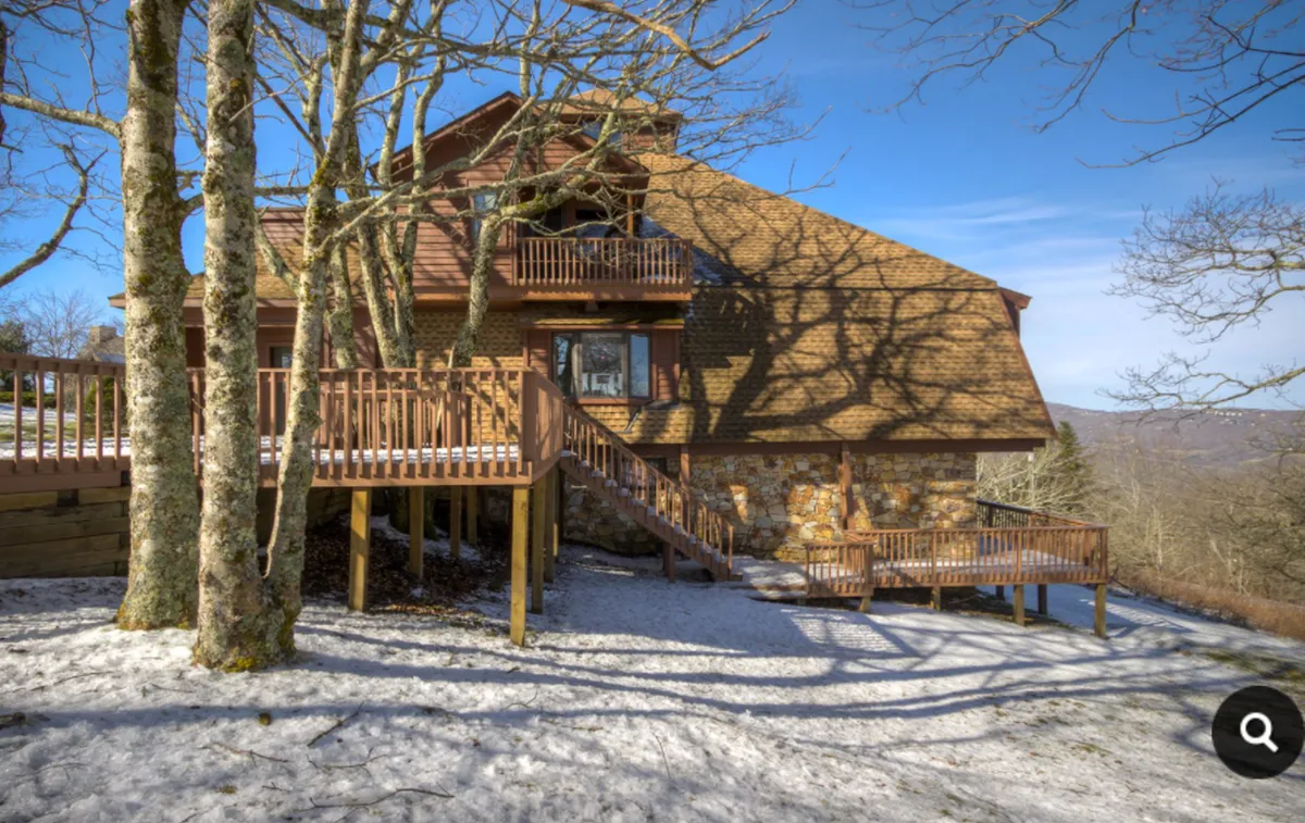 The image captures a wooden deck bathed in tree shadows with a rustic stone wall, inviting guests to relax outdoors or host an intimate gathering. The tranquil surroundings hint at the opportunity for nature walks and quiet moments with friends or family.