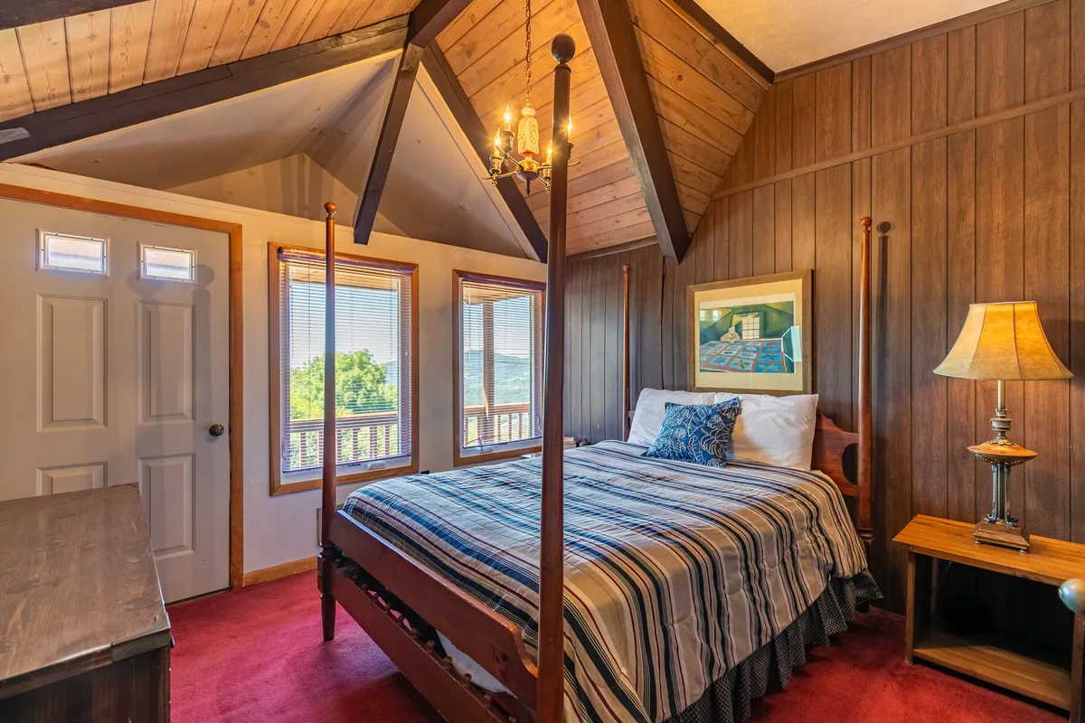 This inviting bedroom features a four-poster king bed dressed in soft linens, a decorative lamp, windows offering scenic views, and warm wood-paneled walls. The space exudes tranquility and comfort, making it an ideal master retreat for rest after adventurous days.