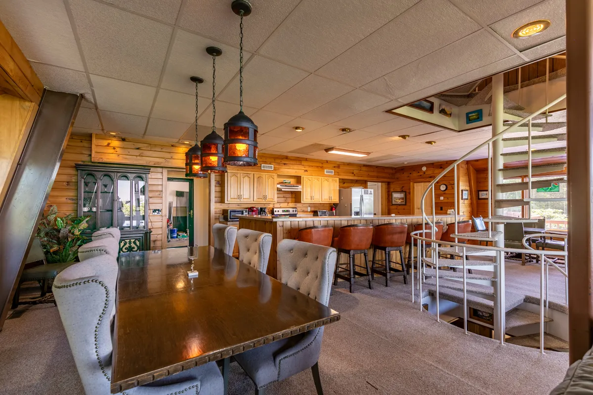 A warm, inviting dining space featuring a wooden dining table, a casual bar area, a spiral staircase, and hanging light fixtures. The area is bathed in natural light, ideal for memorable meals, game nights, or brunches, and seamlessly connects with the rest of the open living space.